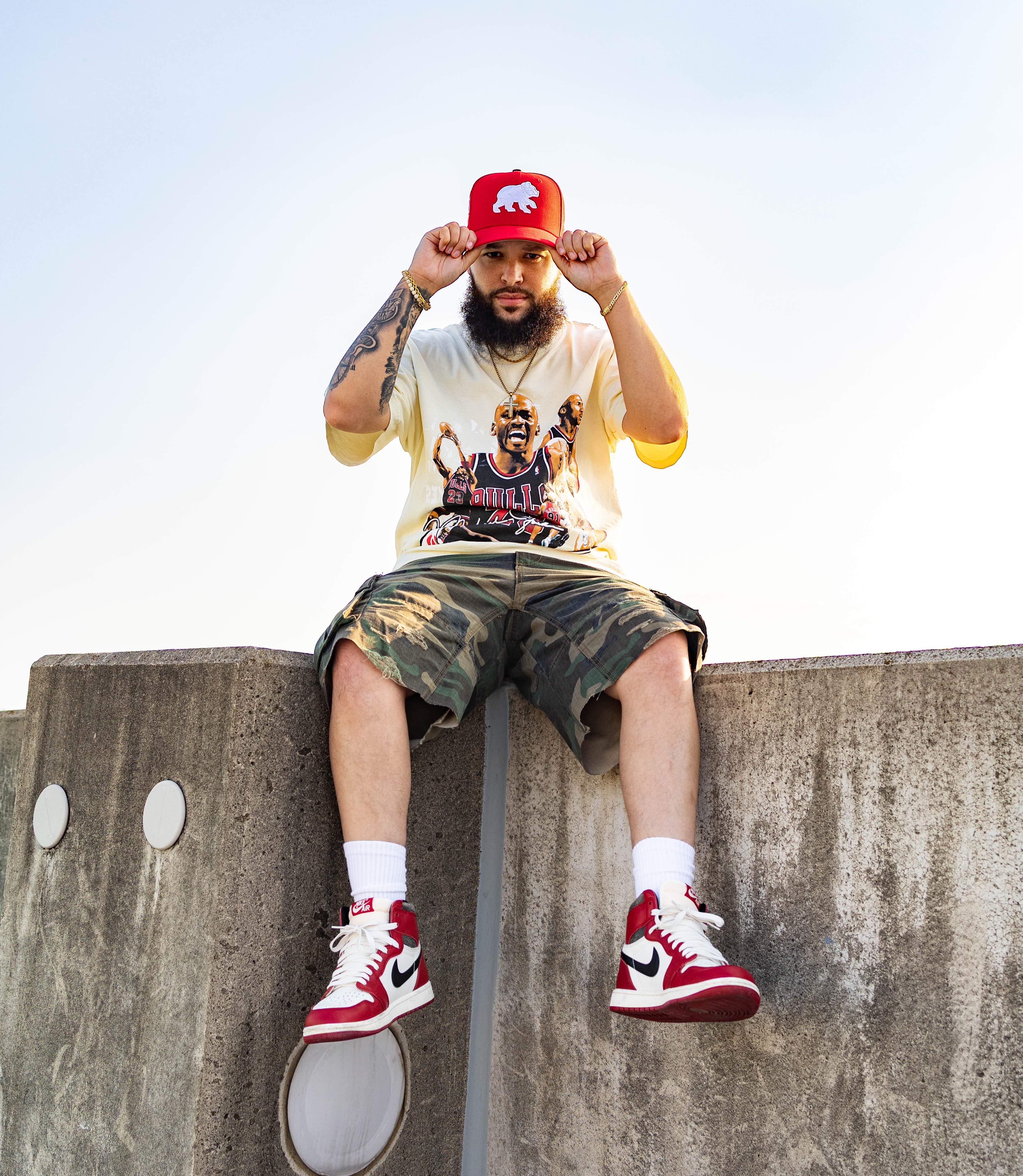 Person sitting on a concrete ledge wearing a red cap, white t-shirt with graphic design, camouflage shorts, and red sneakers.